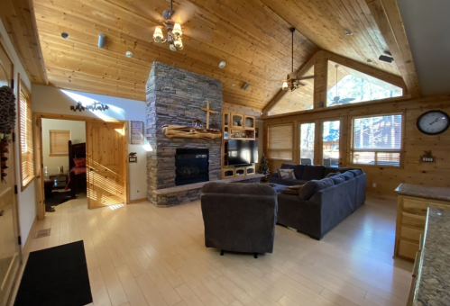 Cozy living room with wooden walls, stone fireplace, large windows, and comfortable seating. Bright and inviting atmosphere.